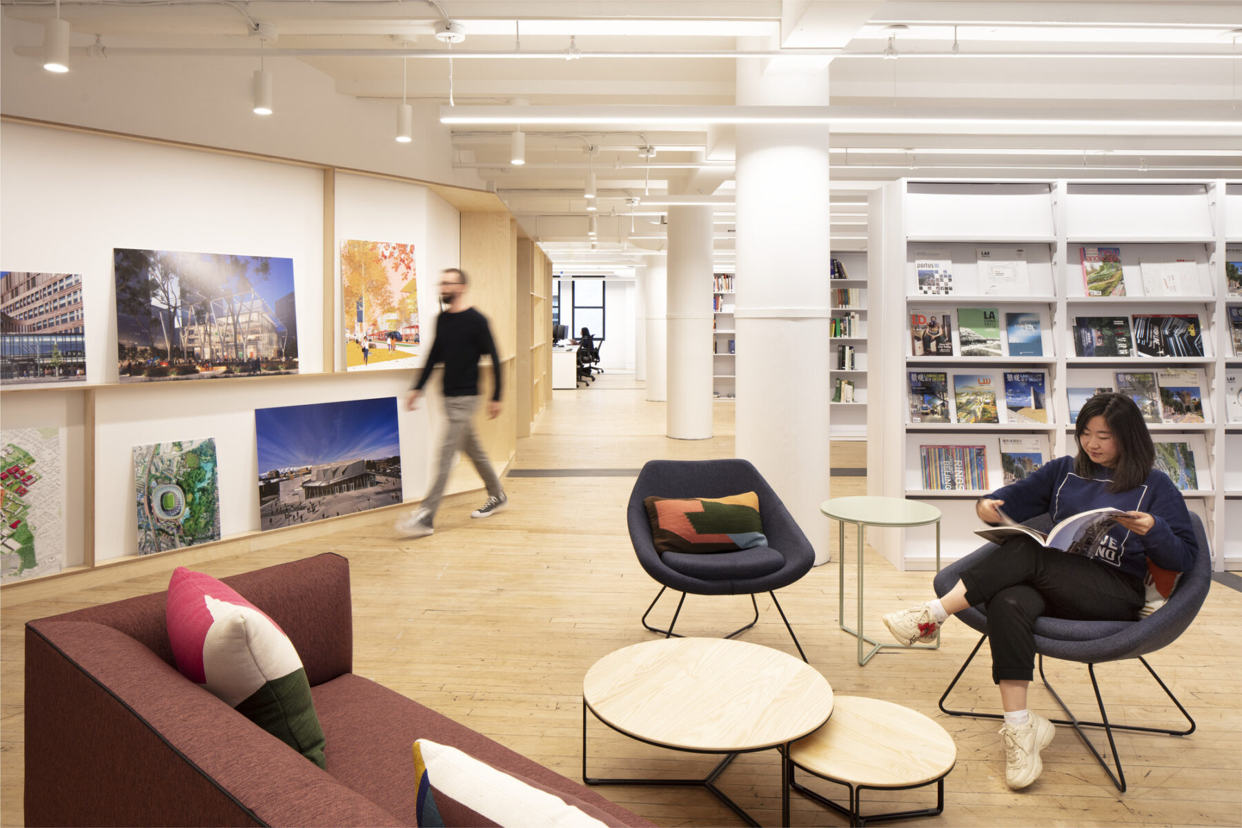 woman reading a magazine in the library with stacks in background and person walking by