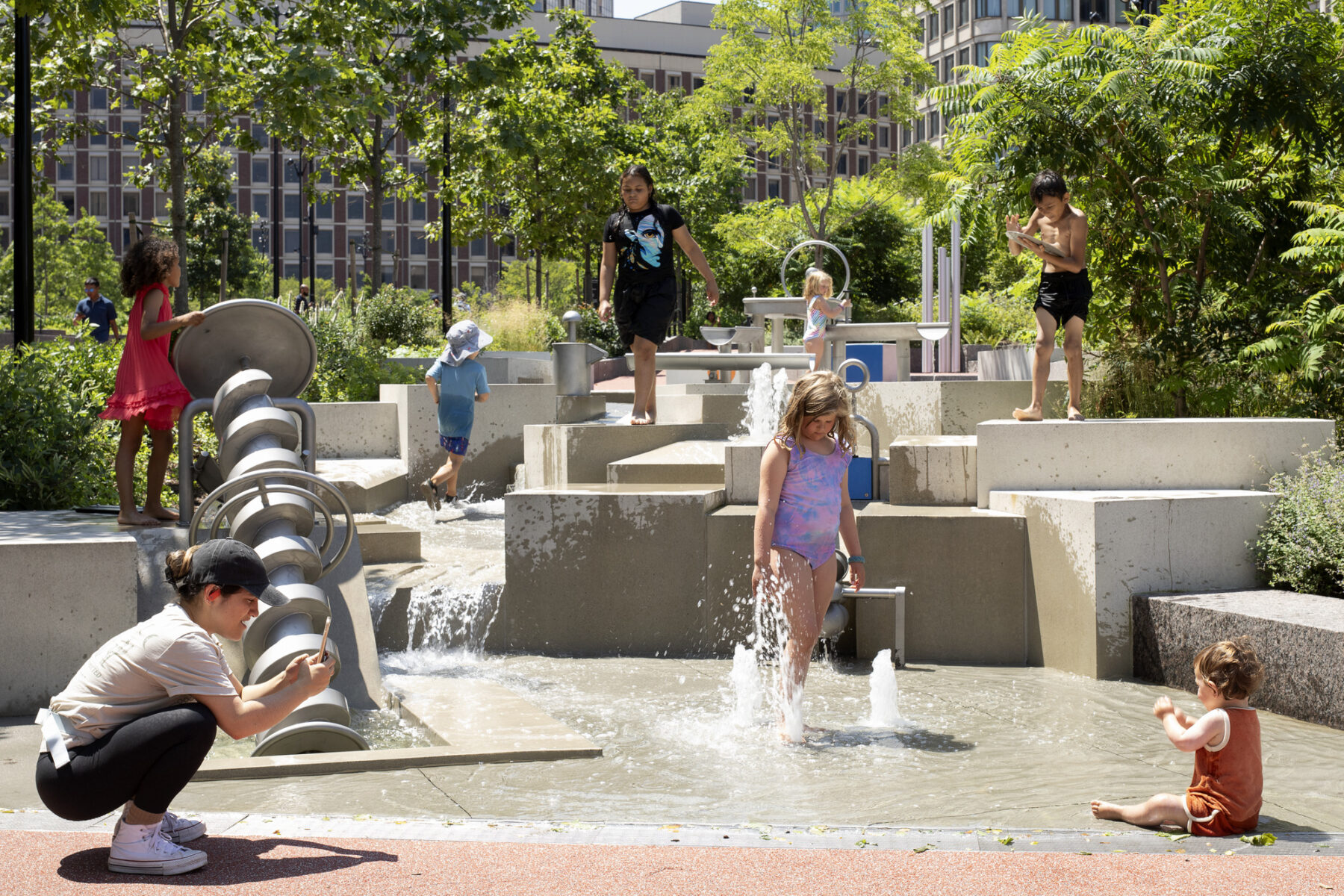 Children climbing on the sculptural water play feature, and a mother in foreground taking a photo of her baby splashing in the shallow water area.