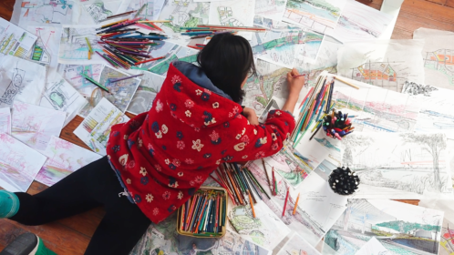 A person lying on the ground drawing, surrounded by drawings and colored pencils