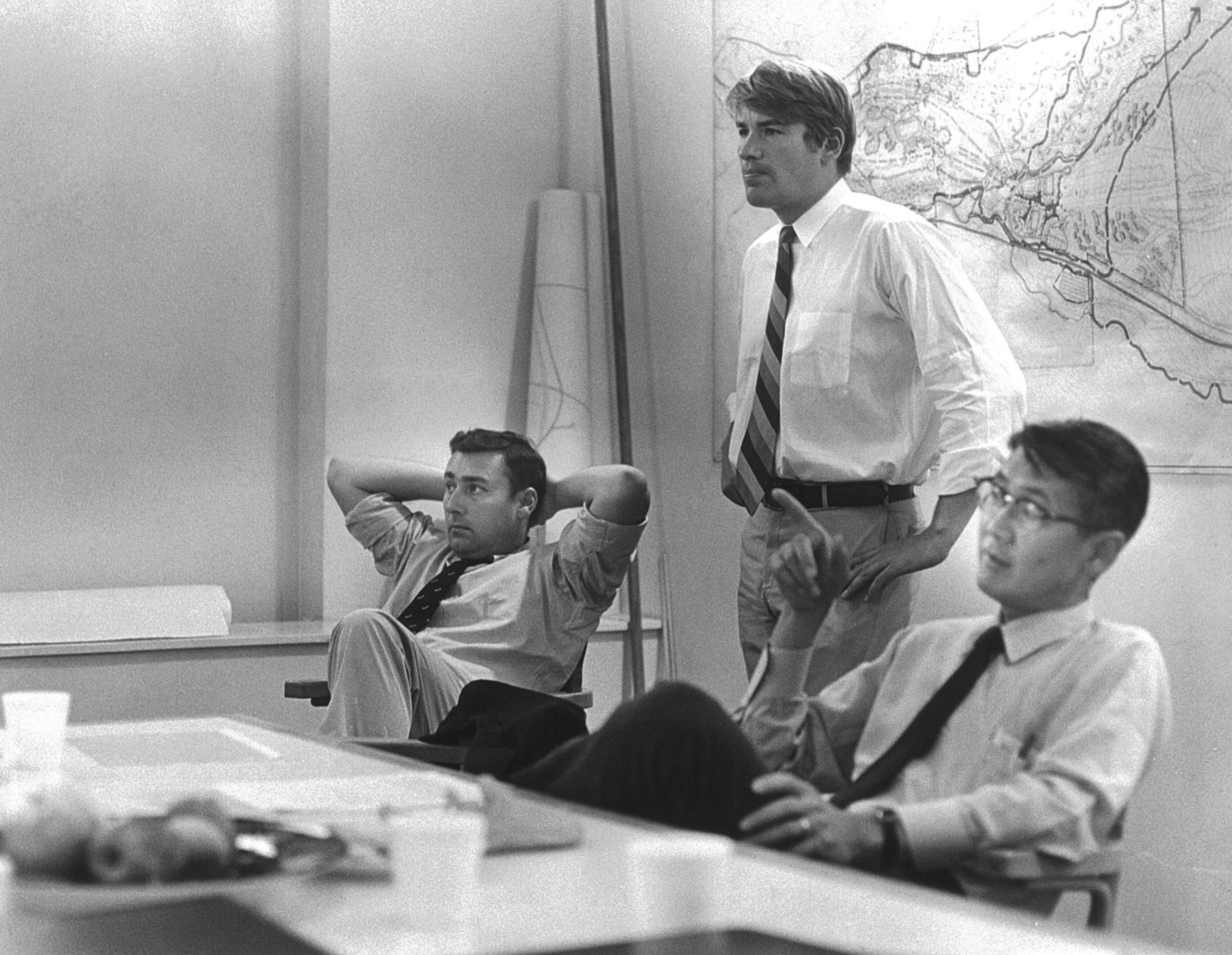 Black and white photo of Hideo Sasaki at a table with two other men. A plan is hanging on the wall in the background